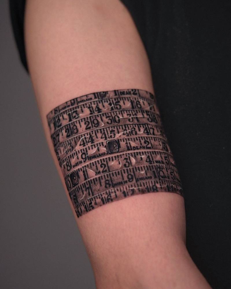 Tape measure tattoo located on the upper arm, black an grey style.