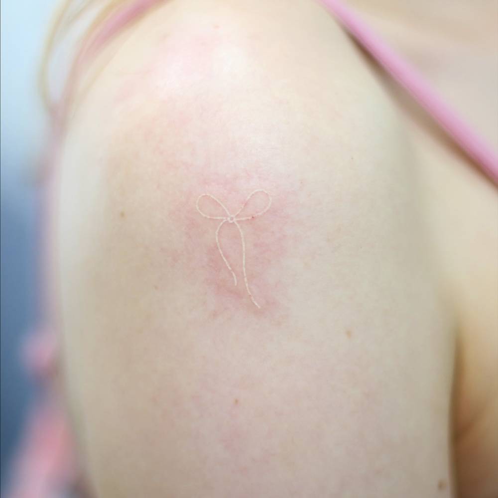 White ink bow tattoo located on the shoulder, minimalistic style.