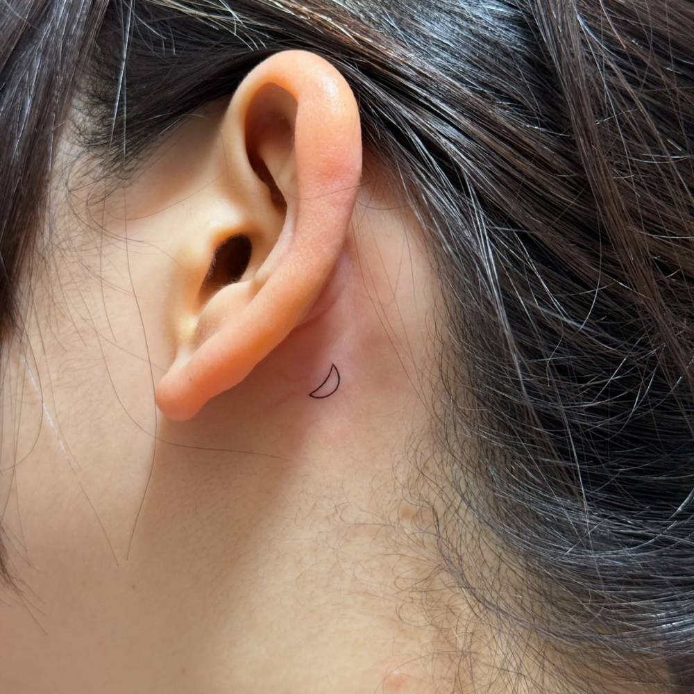 Tiny crescent moon in minimalistic style tattooed behind the ear.