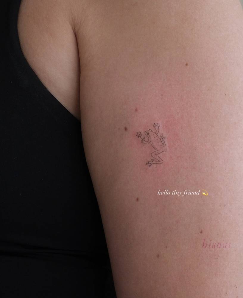 Tiny frog tattoo in fine line style located on the upper arm.