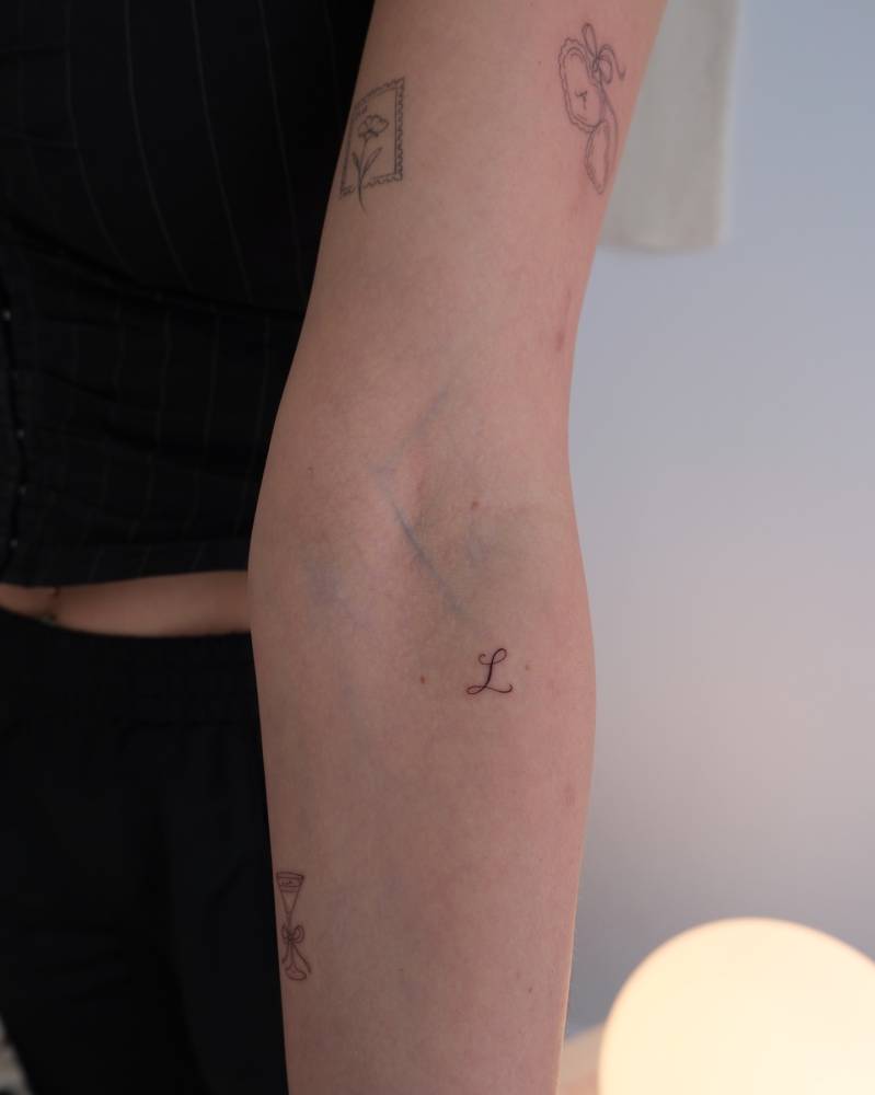 Little letter "L" tattoo in script font located on the inner forearm.