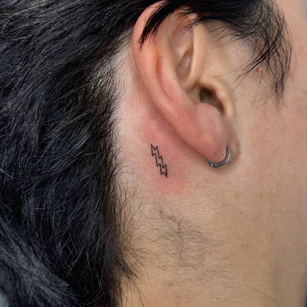 Lightning bolt tattoo in minimalistic style located behind the ear.