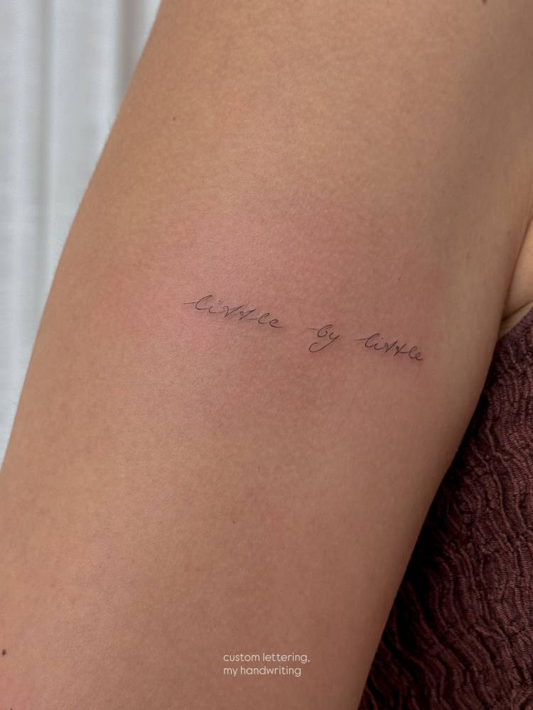 Handwritten font "little by little" tattoo located on the upper arm.