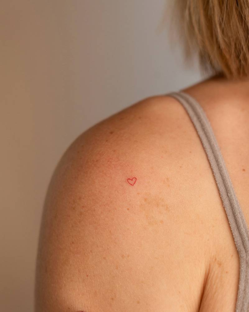 Tiny red heart tattoo in minimalistic style done on the shoulder.