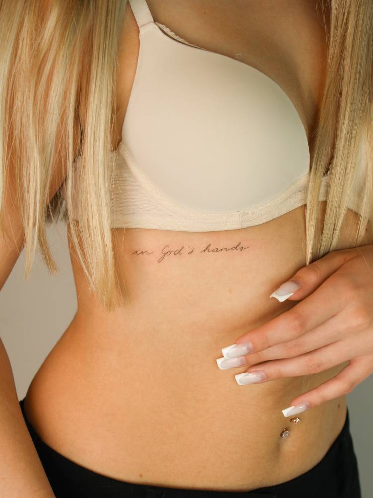 Handwritten font "in God's hands" lettering tattoo located on the rib.