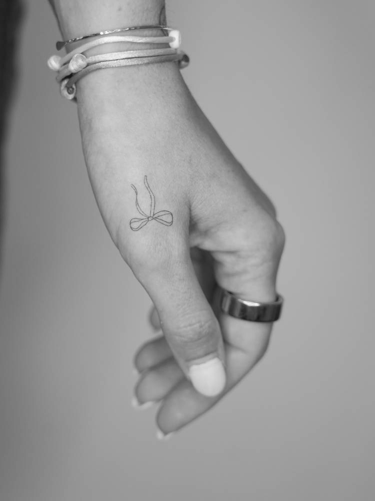 Tiny bow tattoo located on the hand, done in fine line style.