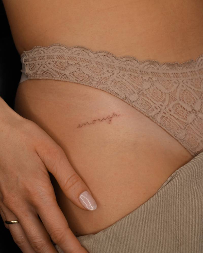 Handwritten lettering tattoo of the word "enough" located on the pelvis.