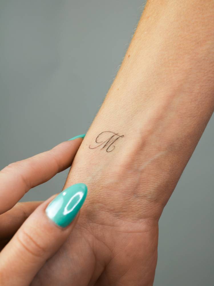 Script font letter "M" tattoo placed on the wrist.