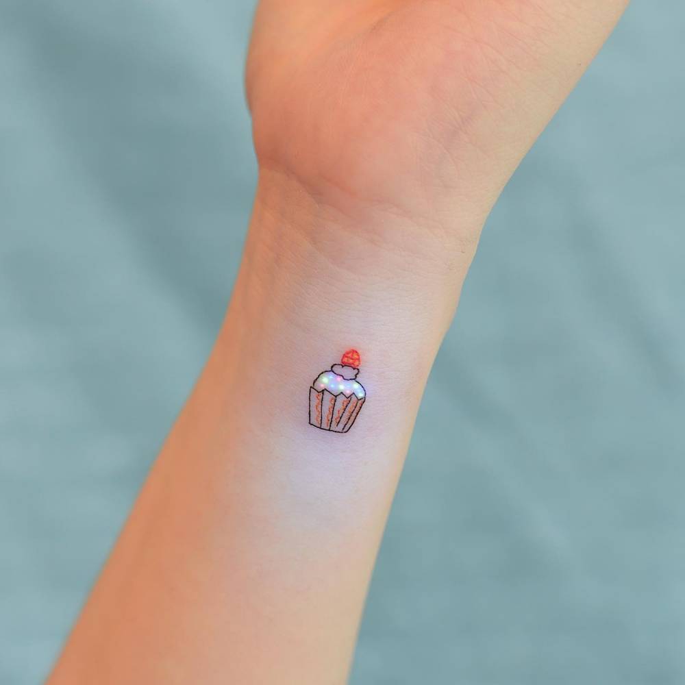 Illustrative style UV ink cupcake tattoo located on the wrist.