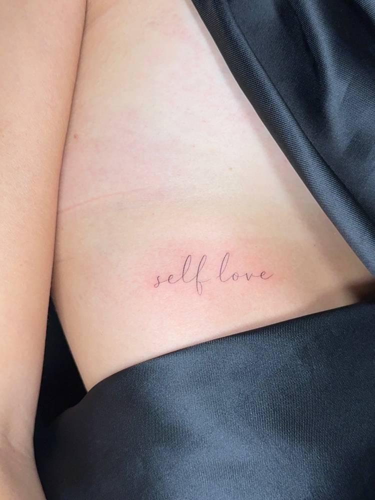 Handwritten font "self love" lettering tattoo located on the rib.