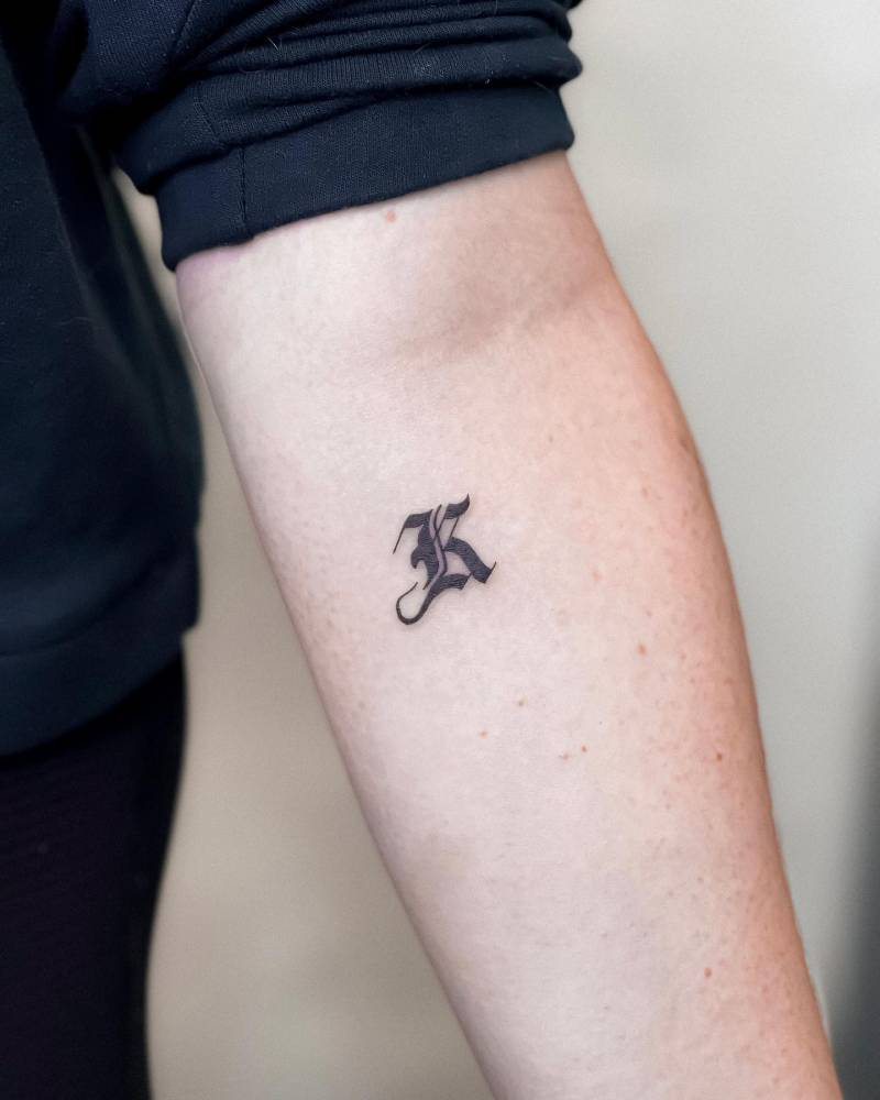 Letter "K" tattoo in gothic font located on the inner forearm.