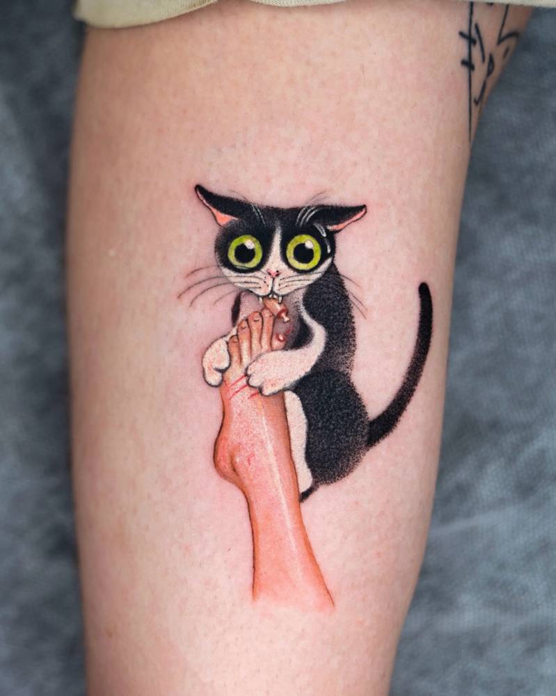 Cartoon style tuxedo cat eating a toe tattooed on the inner arm.
