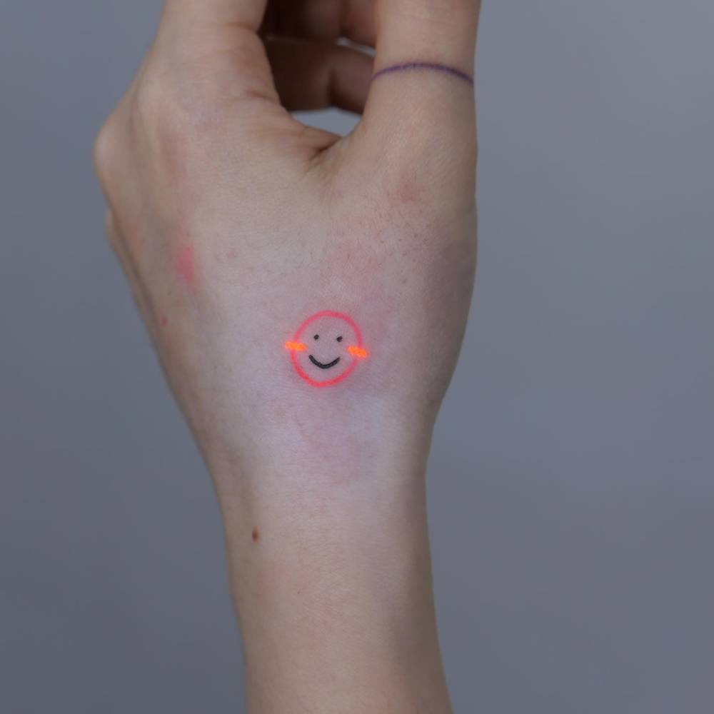UY ink smiley face tattoo in minimalistic style located on the hand.