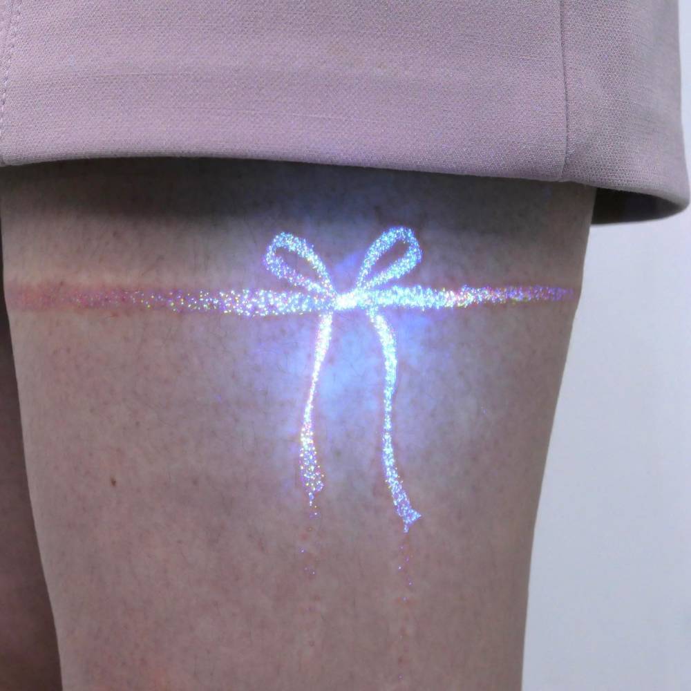 UV ink glitter bow tattoo located on the thigh.