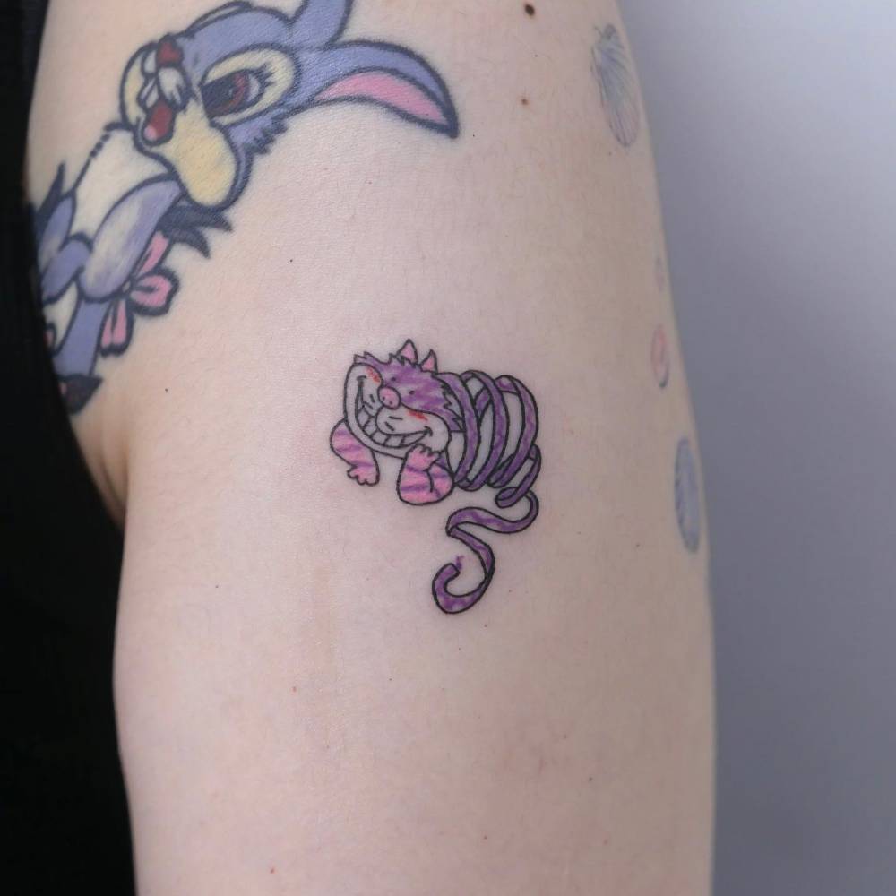 Cartoon style Cheshire Cat tattoo located on the upper arm.