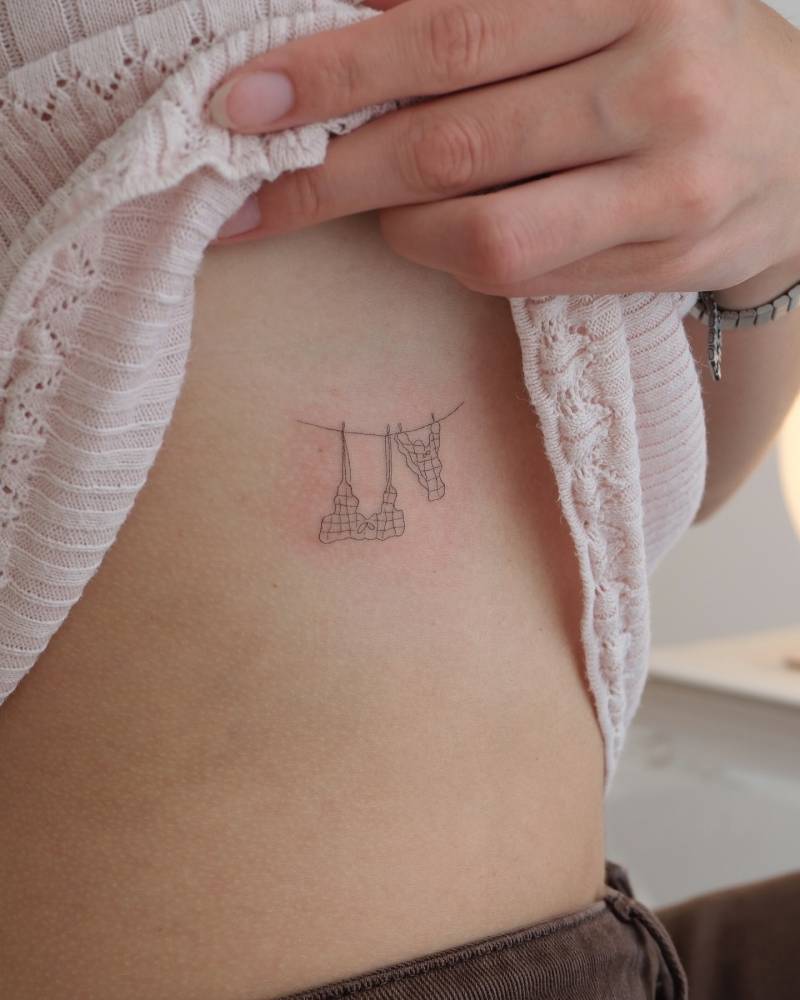 Fine line style bikini hanging on a clothes line tattoo located on the rib.