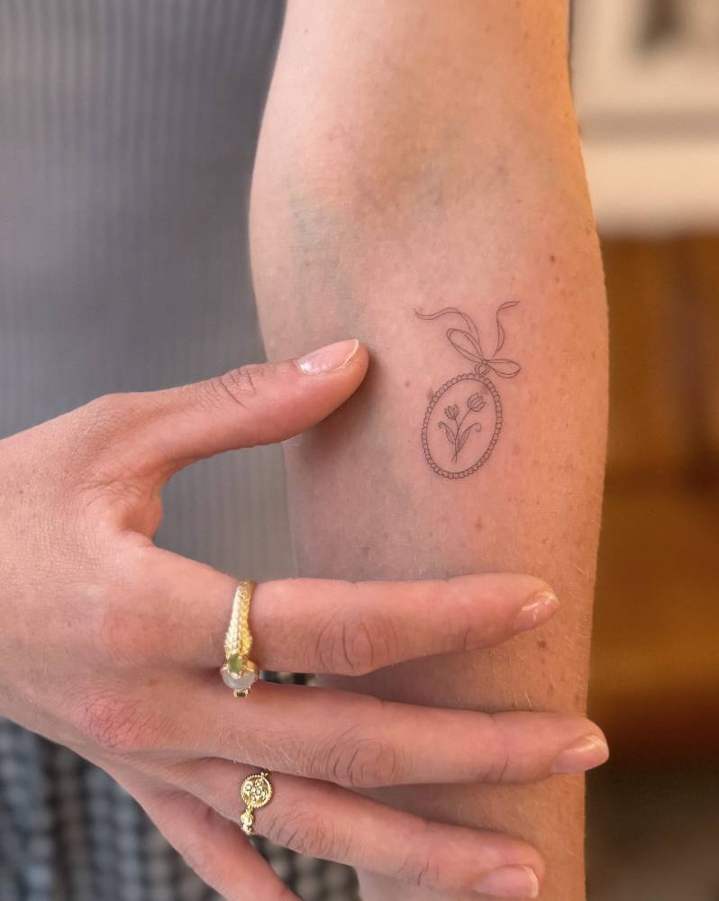 Flower pendant tattoo located on the inner forearm, fine line style.