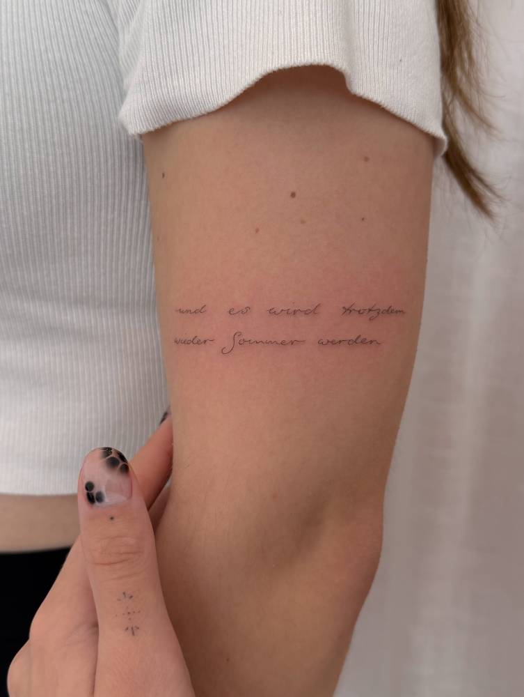 German quote tattoo in handwritten font placed on the upper arm.