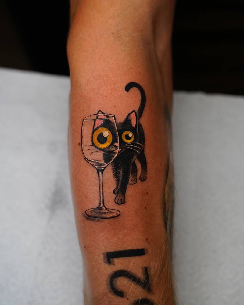 Cartoon style black cat and wine glass tattoo located on the forearm.