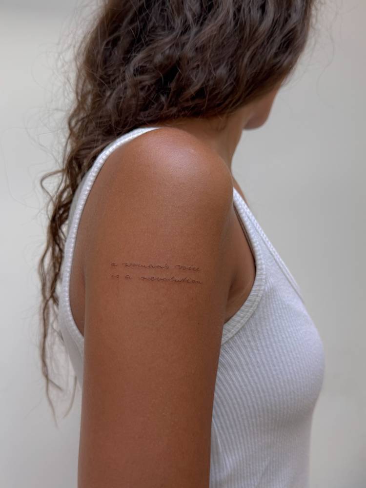 "A woman's voice is a revolution" lettering tattoo placed on the upper arm.