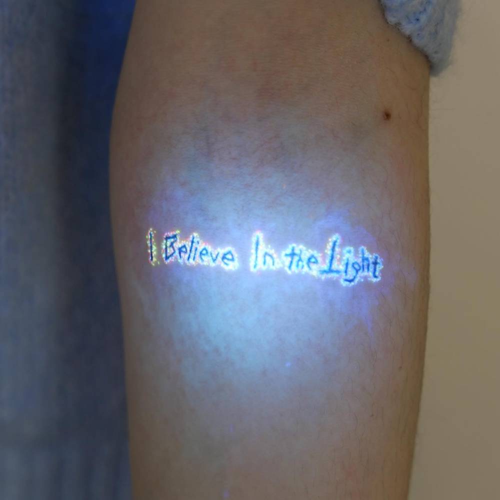 "I believe in the light" lettering tattoo in UV ink located on the inner forearm.