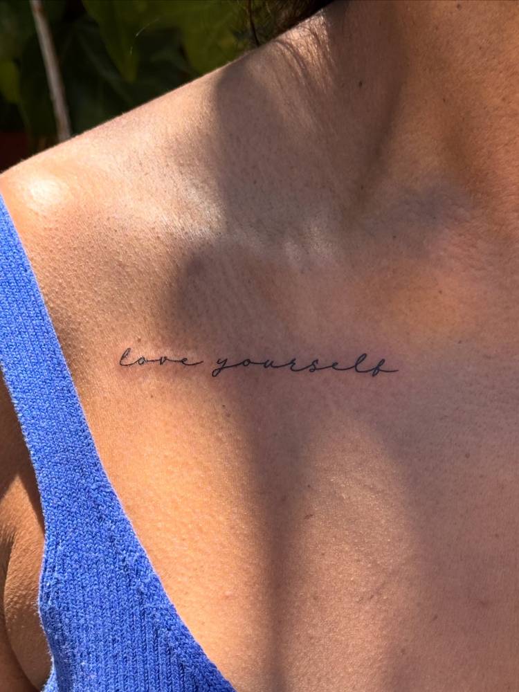 "Love yourself" lettering tattoo in handwritten font located on the collarbone.