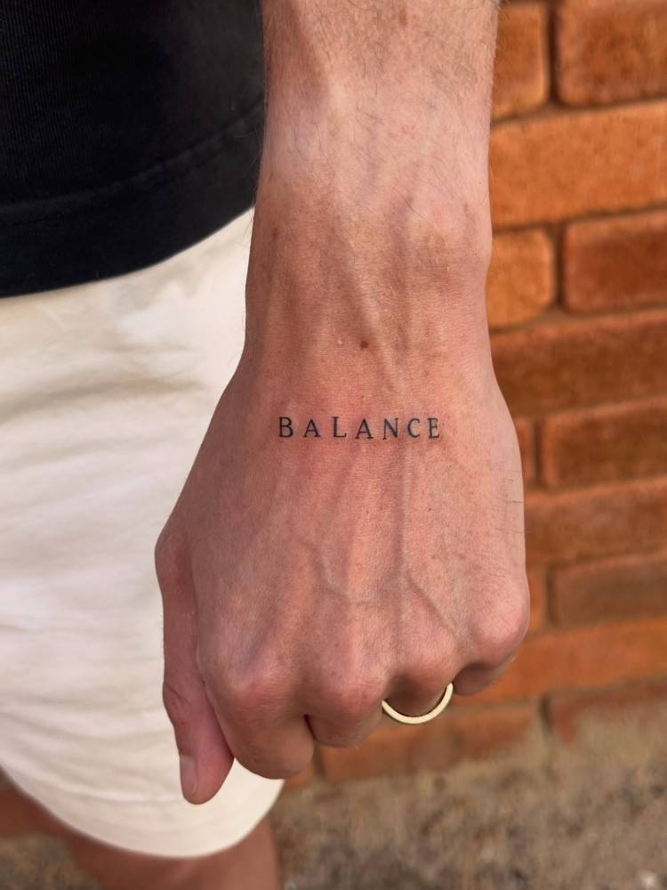 Serif font word "balance" tattooed on the hand.