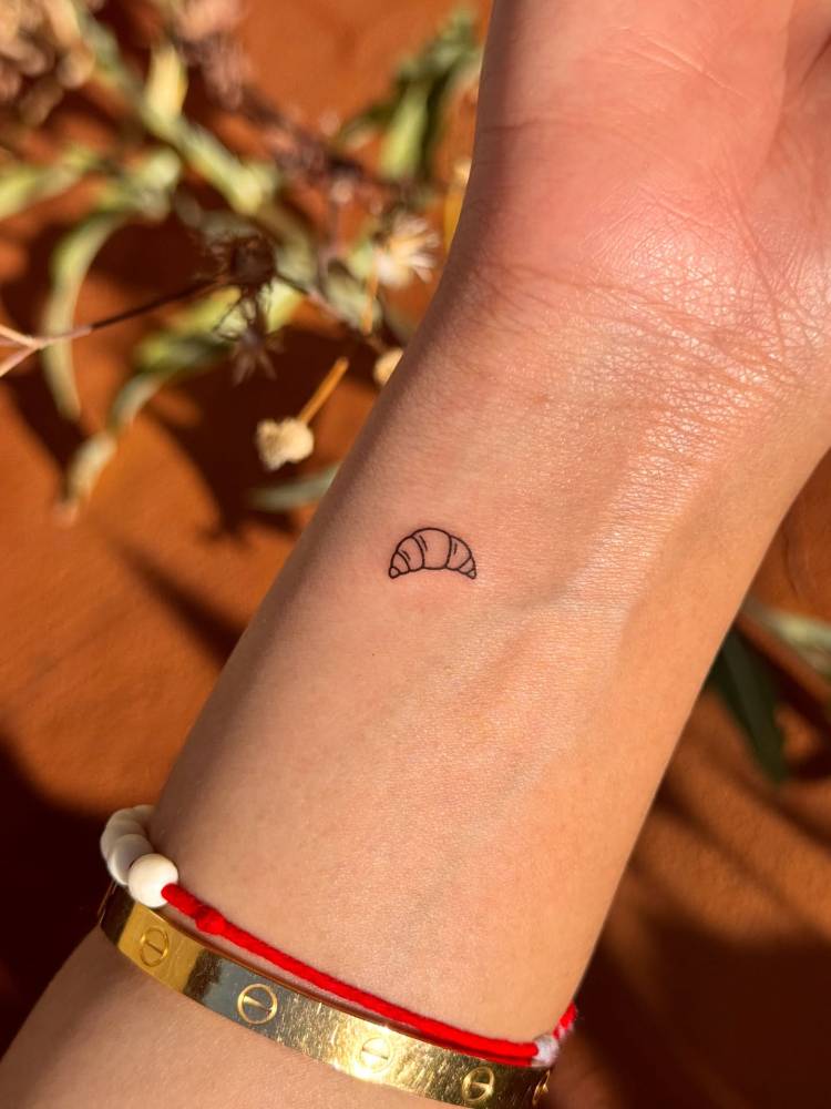 Tiny minimalistic croissant tattoo located on the wrist.