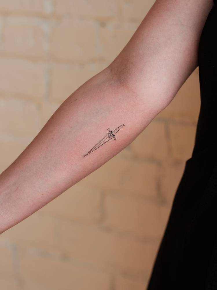 Small dagger tattoo in fine line style done on the inner forearm.