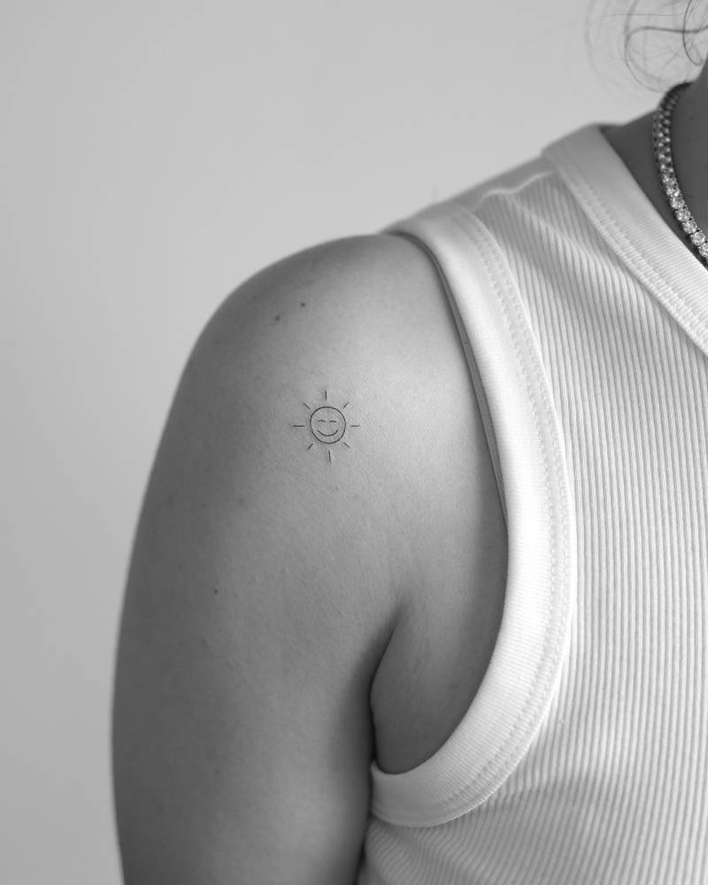 Smiley sun tattoo in minimalistic style located on the shoulder.