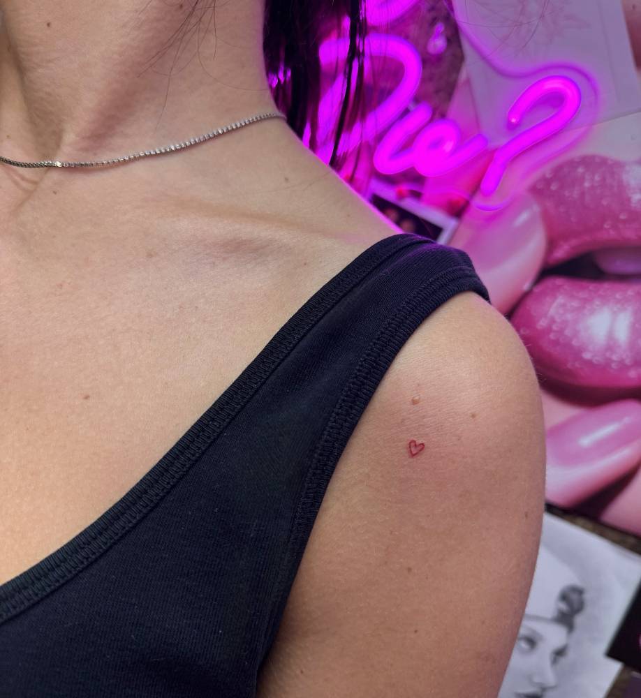 Micro red heart tattoo in minimalistic style located on the shoulder.