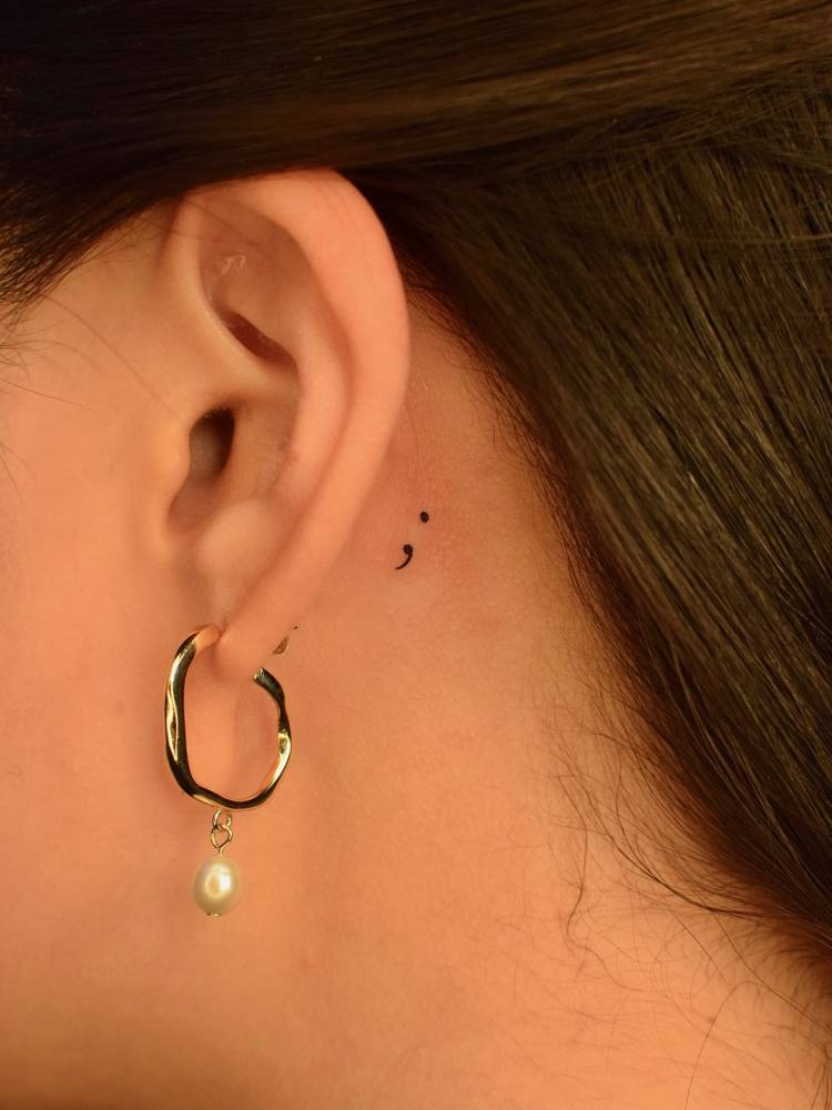 Semicolon tattoo located behind the ear, minimalistic style.