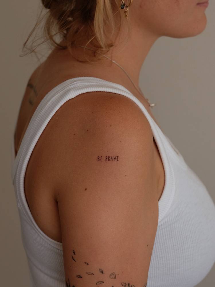 Handwritten font lettering tattoo that says "be brave" placed on the shoulder.