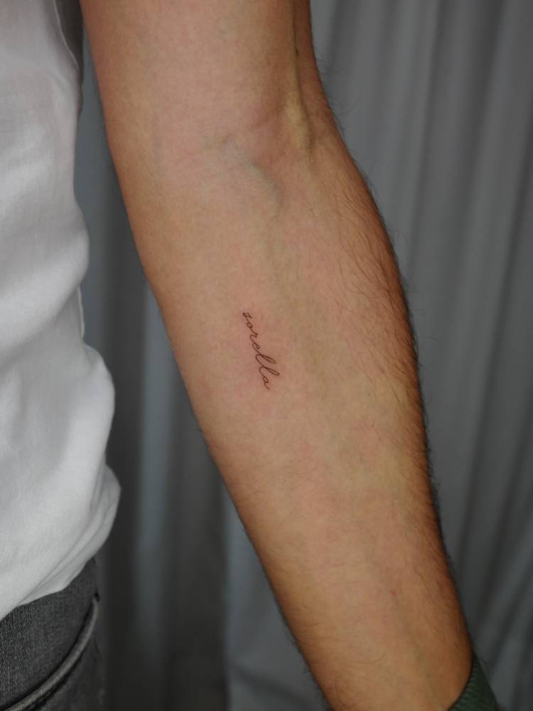 Word "sorella" in handwritten font tattooed on the inner forearm.