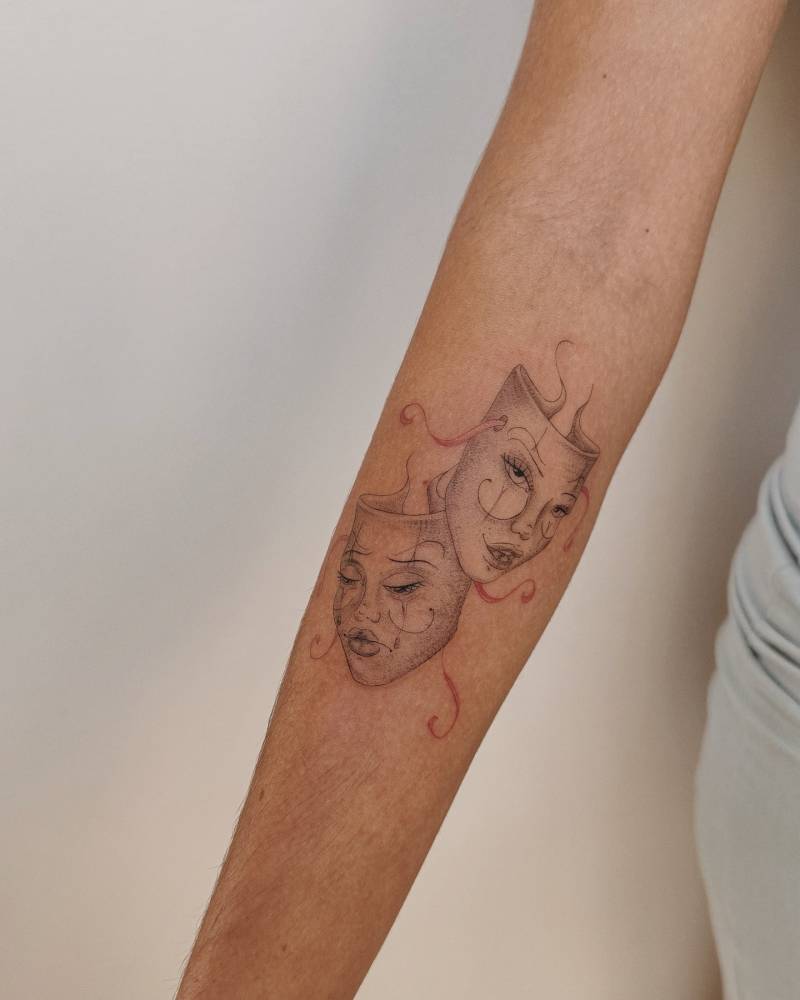 Drama masks tattoo located on the inner forearm, illustrative style.