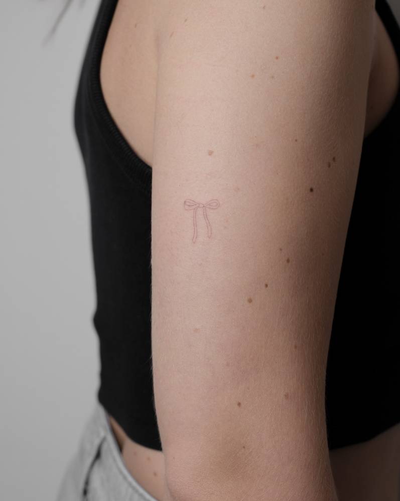 Fine line bow tattoo located on the upper arm, minimalistic style.