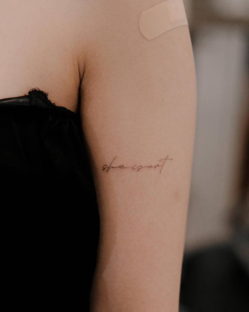 Lettering tattoo that says "she is art" located on the upper arm.