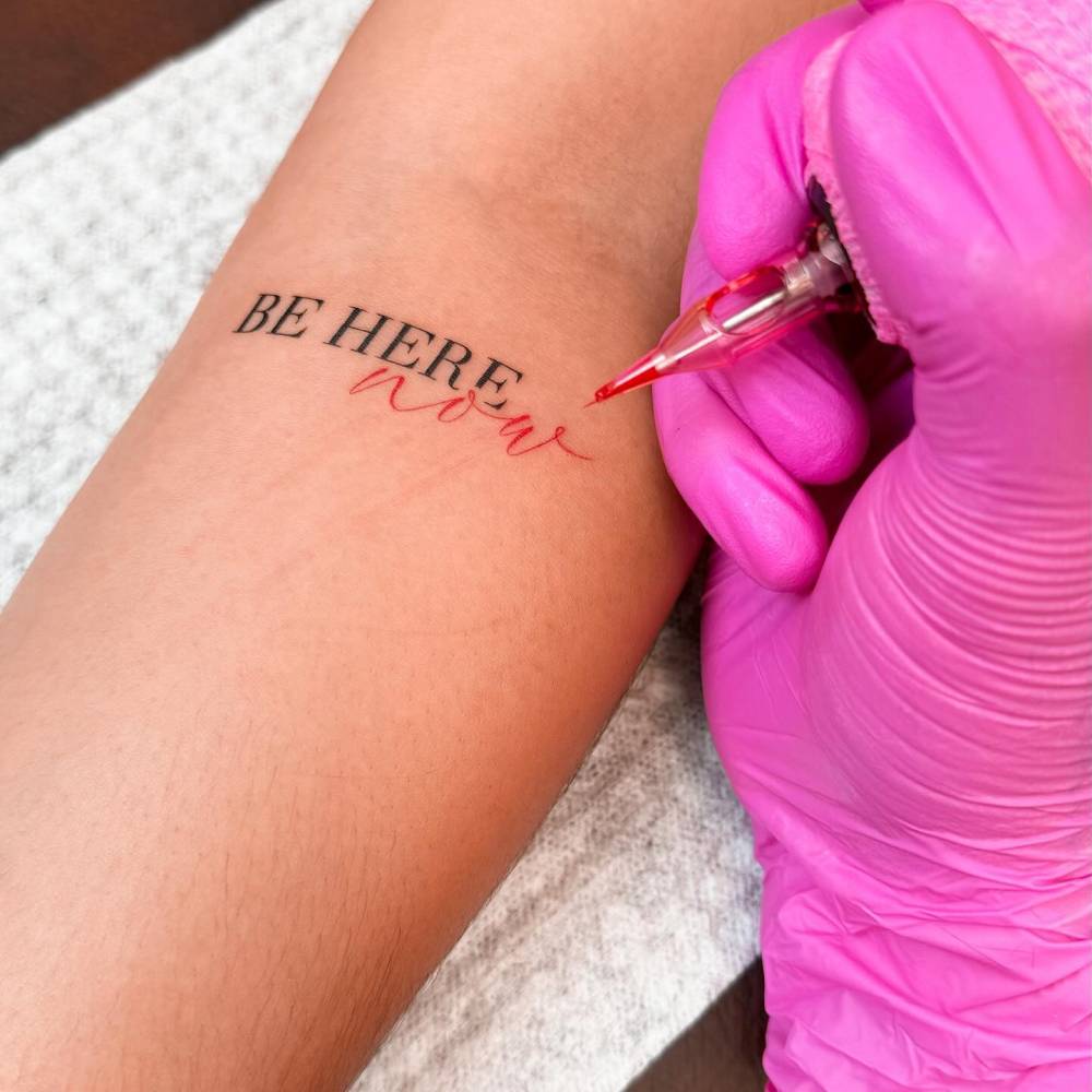 "Be Here Now" lettering tattoo located on the inner forearm.