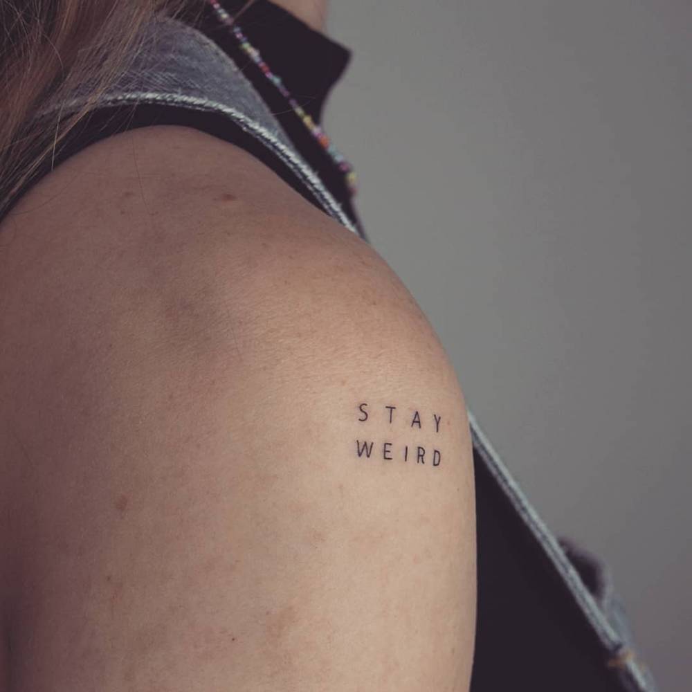 Lettering tattoo that says "stay weird" located on the shoulder.