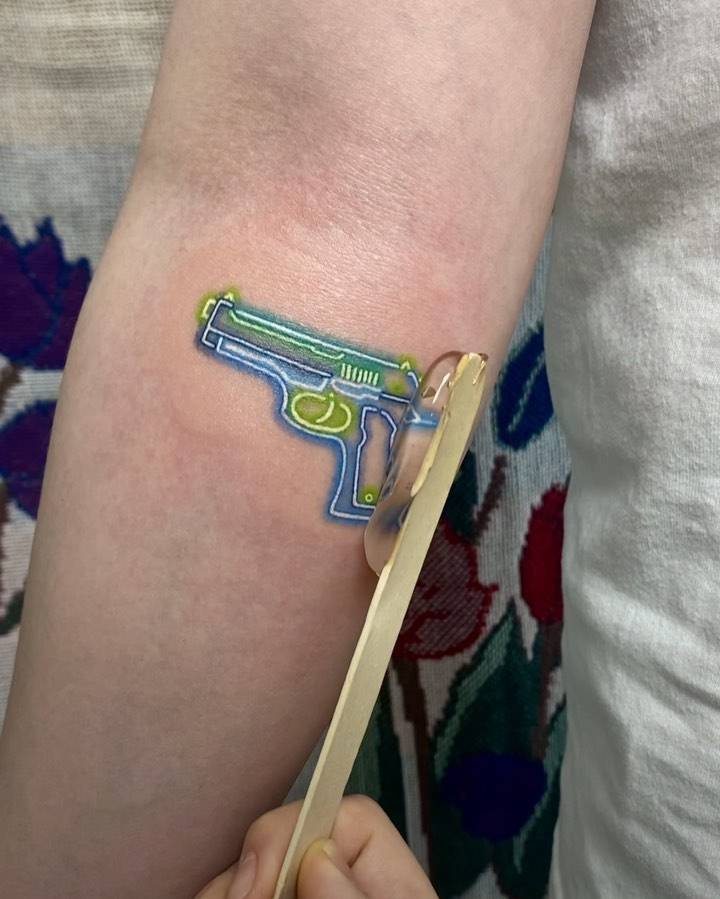 Neon pistol tattoo located on the inner forearm, illustrative style.