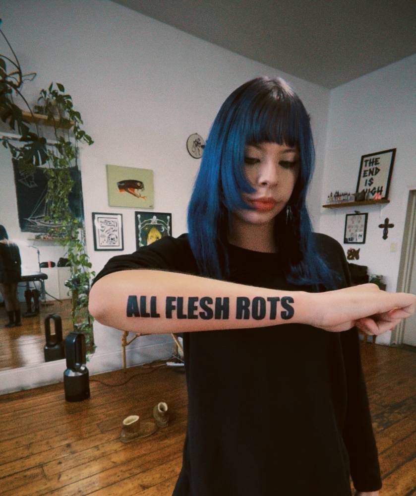 "All flesh rots" lettering tattoo located on the forearm.