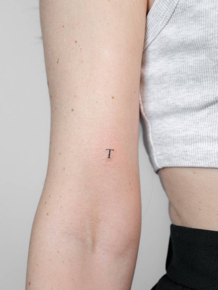 Minimalistic style letter "T" in serif font tattooed on the bicep.