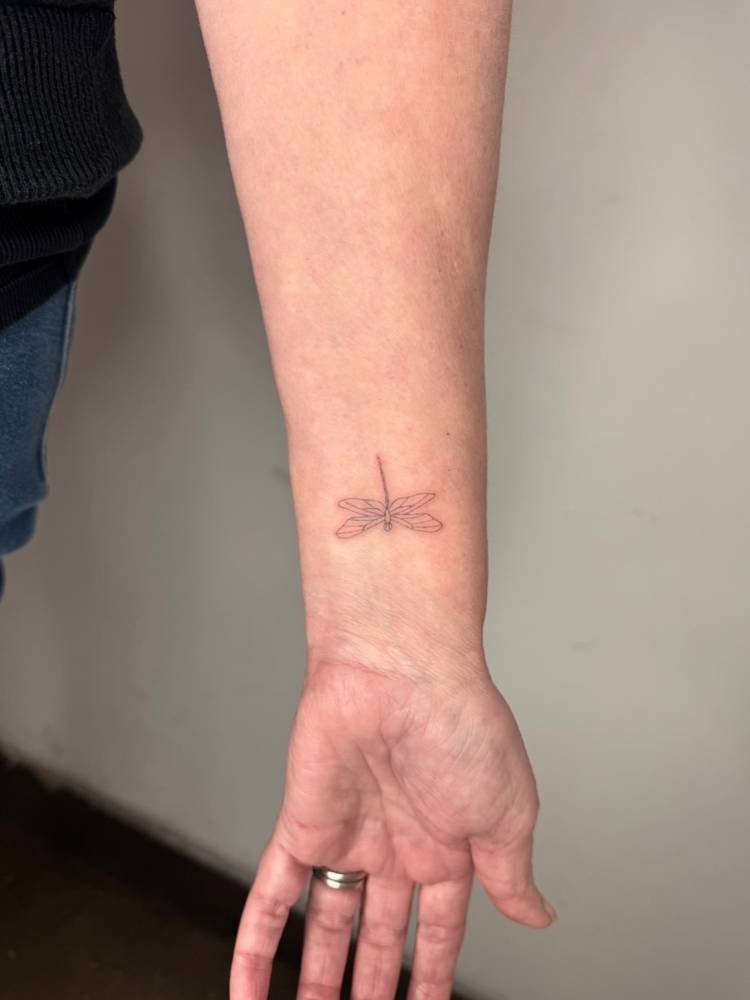 Cute dragonfly tattoo in fine line style placed on the wrist.