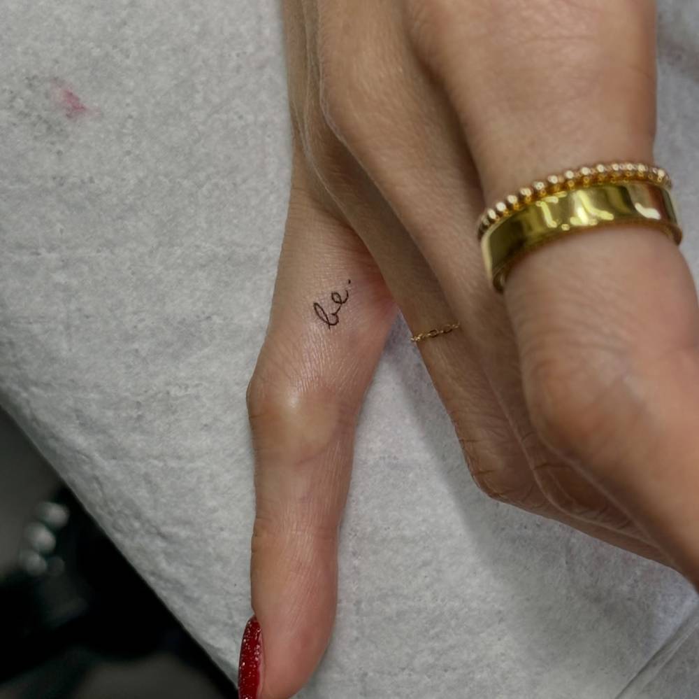 Handwritten font word "be" tattooed on the finger.