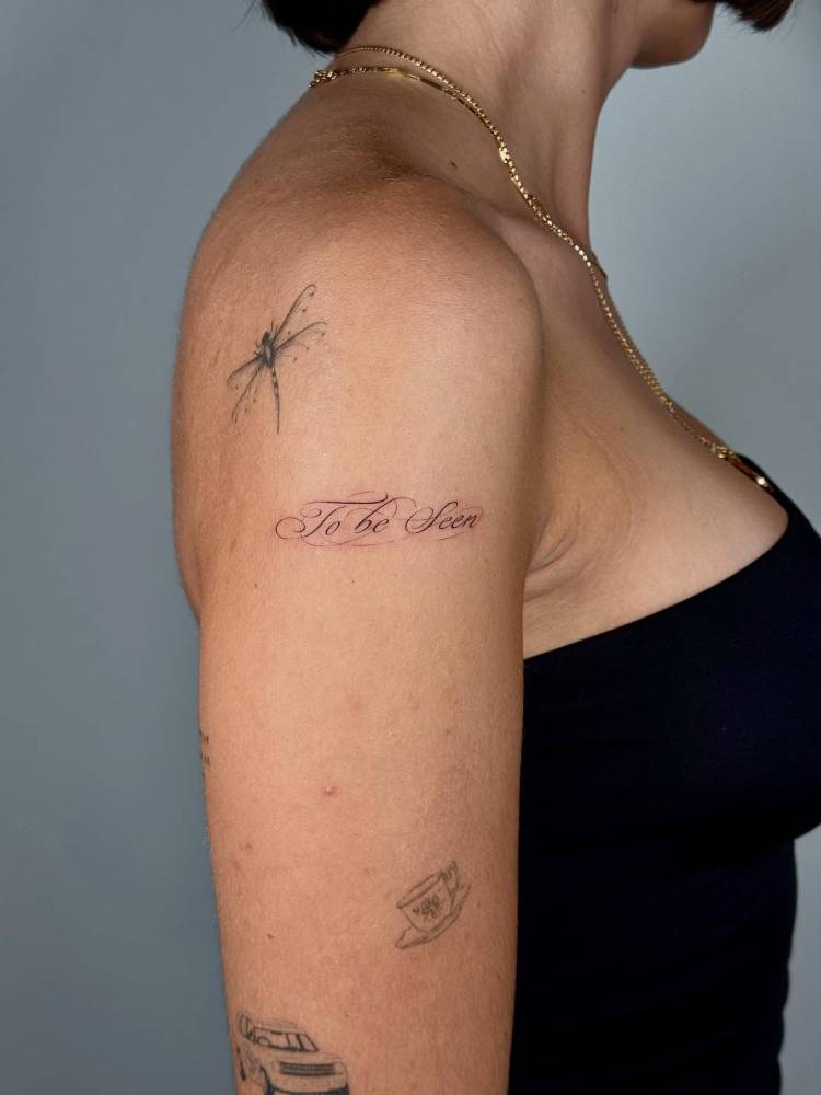 "To be seen" lettering tattoo located on the upper arm.