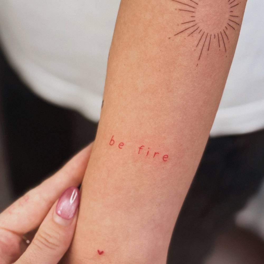 "Be fire" lettering tattoo in red ink located on the forearm.