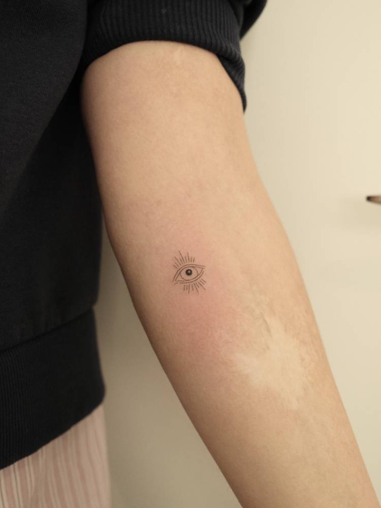Tiny evil eye tattoo in fine line style located on the inner forearm.