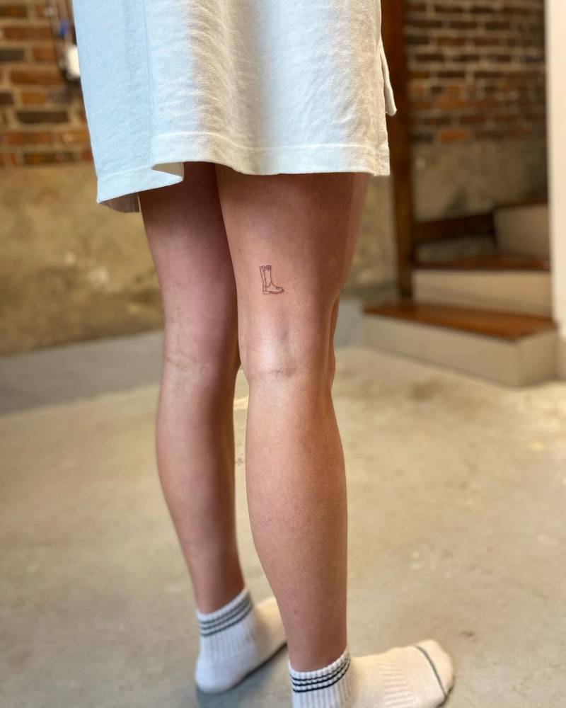 Teeny tiny cowboy boot tattoo located on the thigh, fine line style.