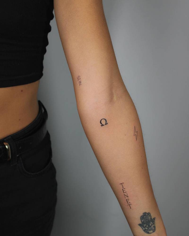 Omega symbol, "Tenet" and lightning bolt tattoos located on the inner forearm.