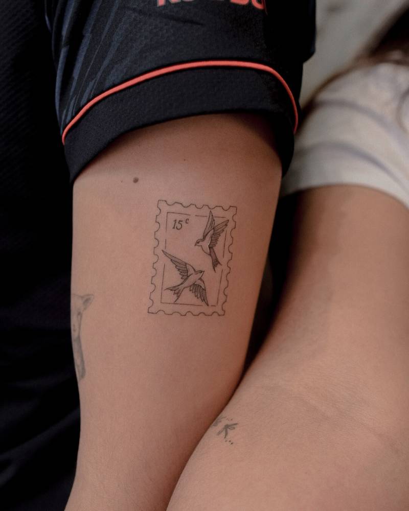 Fine line style swallow stamp tattoo located on the upper arm.